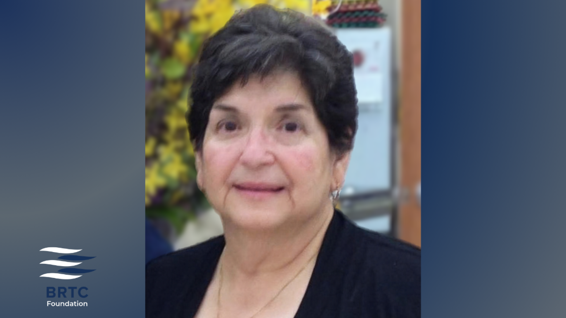 An older woman with short dark hair, wearing a black top, smiles gently in a portrait with flowers blurred in the background, with the BRTC Foundation logo.